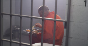 Elderly criminal in orange uniform sits in prison cell eats dinner